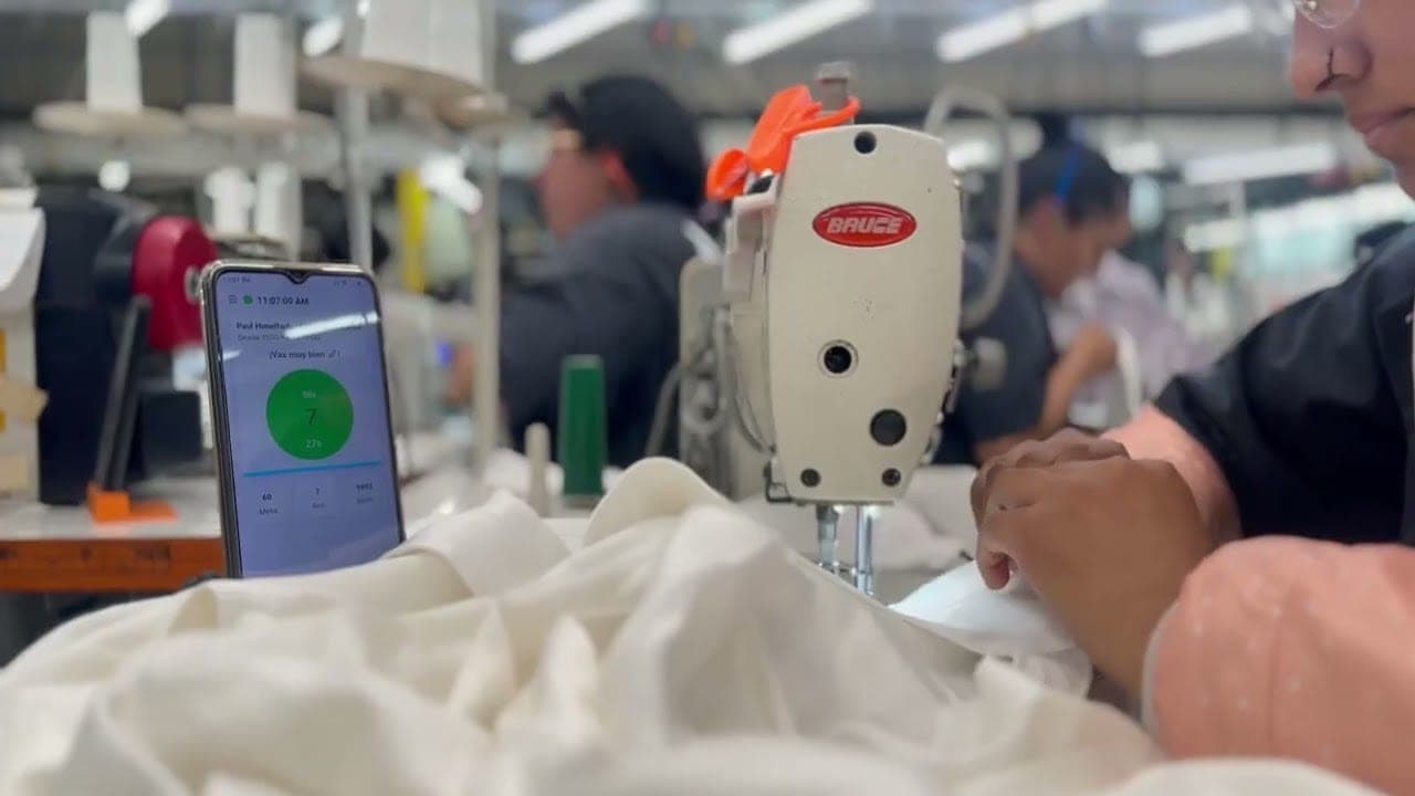 Una planta de confección en plena operación: sobre la máquina, el teléfono del operario marca en verde que va al ritmo de la meta de la hora mientras el módulo sigue cosiendo.